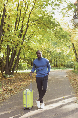 Cheerful african american young man in stylish clothes with suitcase walks in an spring park on warm sunny day. Countryside travel conceptの写真素材