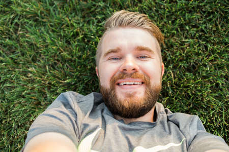 young man with beard and fashion hair style lying on grass taking selfie - holding smart phone or...