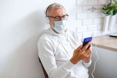 Senior man with medical mask on face using smartphone app for creating playlist music - Elderly male having fun with mobile phone technology - Tech and joyful elderly lifestyle conceptの写真素材