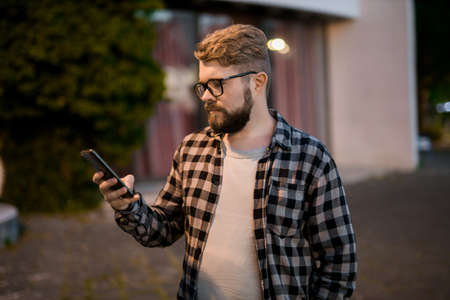 Bearded man wearing eyeglasses is holding scrolling texting in his cellphone at night street. Guy...
