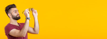 Handsome arabian guy is looking at his glasses while standing against a yellow background banner with copy space and place for advertising. Poor vision and eye treatment conceptの写真素材
