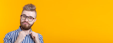 Cute young hipster student guy with a mustache and a beard with glasses straightens his shirt while posing on a yellow background. The concept of self-confidence. Advertising spaceの写真素材