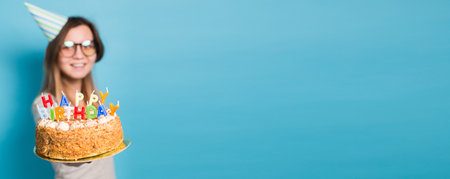 Close up funny positive girl in glasses and greeting paper hat holding a happy birthday cake in her hands standing on a blue background banner with copy space and place for advertisingの写真素材
