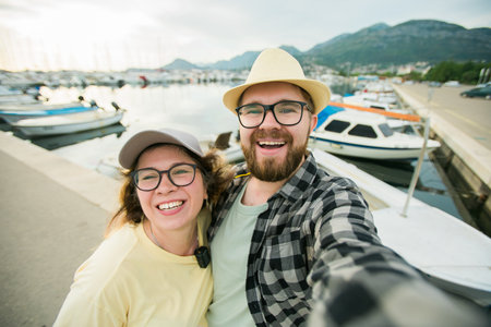 Young couple taking a self portrait laughing as they pose close together for camera on their smartphone outdoors in summer port marina with boats and yachtsの写真素材