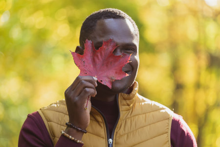 Close up portrait of african american man covering his face with autumn maple leaf. Autumn nature....