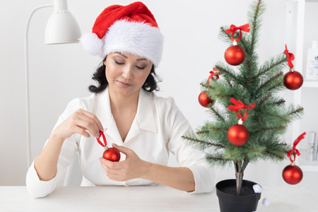 Portrait of smiling latin or indian business woman decorating christmas tree in office. Happy middle eastern woman decorating tree for christmas holiday. Multiethnic people celebrating holiday work.の写真素材