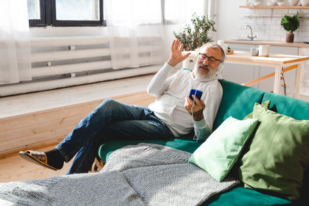 Elderly man cool bearded old man using mobile phone for video call - happiness elderly lifestyle people concept. Video call on smartphone in room waving to screen and chatting with children - modern technologies communication and internet.の写真素材