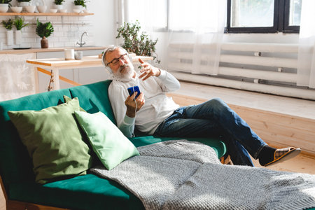 Elderly man cool bearded old man using mobile phone for video call - happiness elderly lifestyle people concept. Video call on smartphone in room waving to screen and chatting with children - modern technologies communication and internet.の写真素材