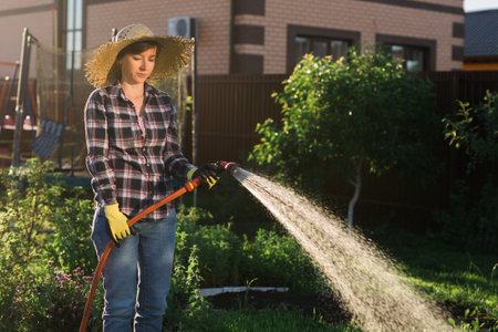 Caucasian woman gardener in work clothes watering the beds in her vegetable garden on sunny warm...