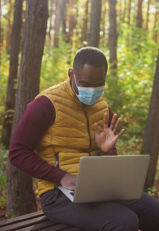 African american man sitting in city park on a bench with laptop studying online outdoors. Man freelancer learn working remotely street e-learningの写真素材