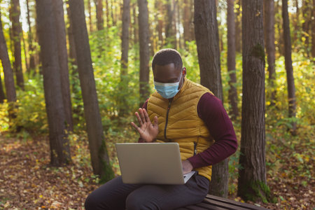 African american man sitting in city park on a bench with laptop studying online outdoors. Man freelancer learn working remotely street e-learningの写真素材