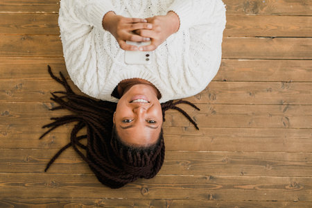 Positive young african american woman chatting on cellphone and smiling on floor at home. Cheerful young woman communicating with someone on smartphone indoorsの写真素材