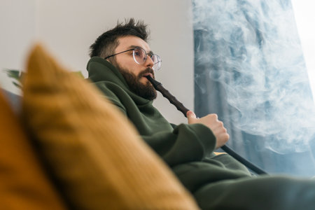 Bearded millennial or gen z man smoking hookah while relaxing on sofa at home - chill time and...