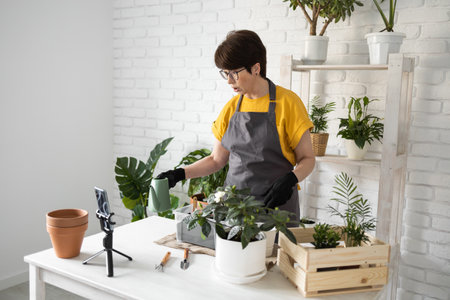 Relaxing home gardening. Smiling middle aged woman in black gloves with potted plant records gardening video blog in modern house - blogging and florist vlog influencer.の写真素材
