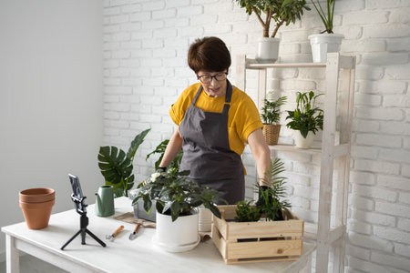 Female blogger sits in front of smartphone camera on tripod records instructional tutorial video for her blog shoots process of replanting flowers and green plantsの写真素材