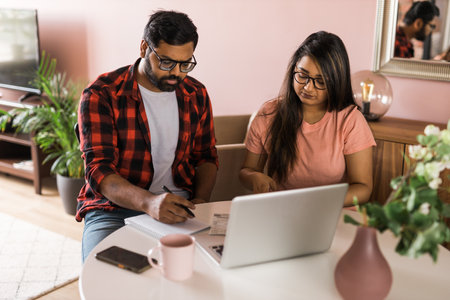 Young millennial indian husband and wife doing domestic paperwork, accounting job and reviewing paper bills, receipts at laptop computer, using online calculator and paying mortgage rent fees on internet - economic crisisの写真素材