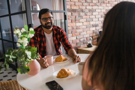 Happy couple eating breakfast and talking at dining table in morning. Indian girl and latino guy. Relationship and diversity conceptの写真素材