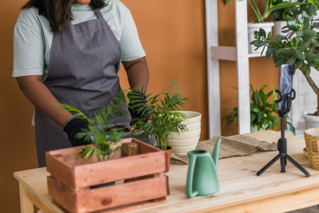 African american girl blogger influencer work on home video camera selfie shoot filming take care home plants and transplanting plant in flowerpot. Home gardening and florist concept.の写真素材