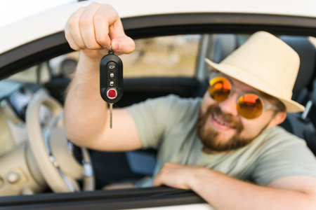 Car driver man smiling showing new car keys and car. Bearded guy driving rented cabrio on summer vacationの写真素材