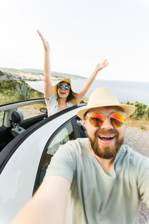 Romantic couple making selfie on smartphone camera in rental cabrio car on ocean or sea beach enjoying summer vacation together and taking picture on cellular resting near sea on weekendsの写真素材