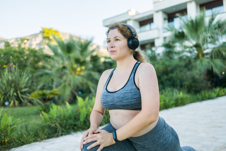 Young pretty smiling plus size woman in sporty top and leggings doing sport and listen to music with headphones in summer morning outdoor copy spaceの写真素材