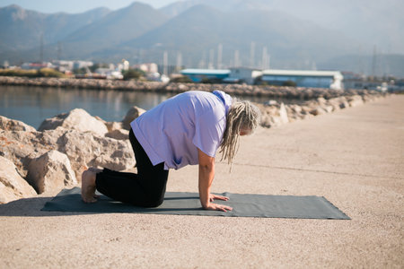 Senior woman with dreadlocks in stretching position by the sea at morning. Elderly woman doing yoga near beach.の写真素材