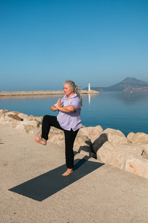 Mature woman with dreadlocks working out doing yoga exercises on sea beach copy space - wellness well-being and active elderly age conceptの写真素材