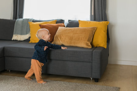 Toddler boy laughing having fun standing near sofa in living room at home copy space. Adorable baby making first steps alone. Happy childhood and child care conceptの写真素材