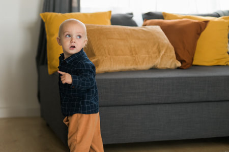 Toddler boy laughing having fun standing near sofa in living room at home copy space. Adorable baby making first steps alone. Happy childhood and child care conceptの写真素材