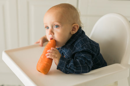 Little boy in a blue t-shirt sitting in a childs chair eating carrot copy space and empty space for text - baby care and infant child feeding conceptの写真素材
