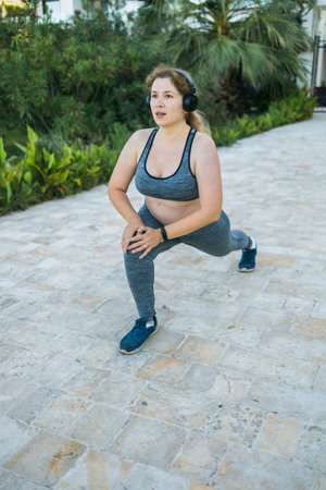 Adorable fat woman in tracksuit is engaged in fitness outdoor side view portrait copy space . Young overweight woman lunges outdoors on warm summer day. Healthy lifestyle and weight lossの写真素材
