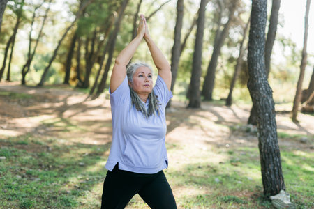 Senior woman with dreadlocks in stretching position on nature at morning. Elderly woman doing yoga...