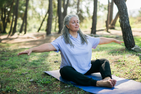 Senior woman with dreadlocks in stretching position on nature at morning. Elderly woman doing yoga on park - wellbeing and wellness tai chiの写真素材