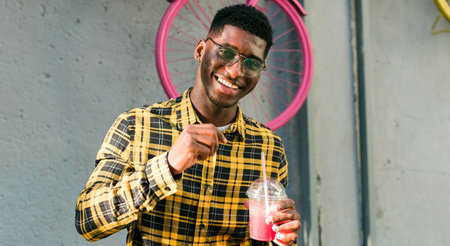 Banner carefree african american guy in casual wear holding tropical cocktail urban summer background copy space. Cool young man drinking yummy alcohol-free beverage on summer vacationの写真素材