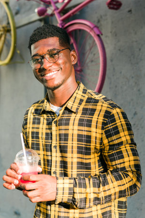 Carefree african american guy in casual wear holding tropical cocktail urban summer background. Cool young man drinking yummy alcohol-free beverage on summer vacationの写真素材