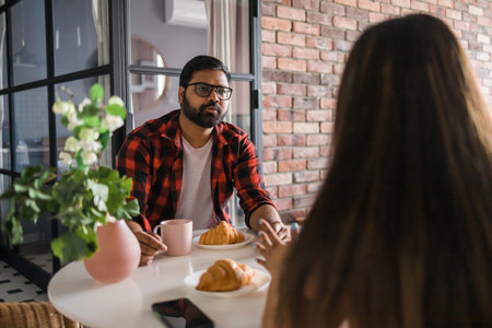 Young diverse loving couple eating croissant and talks together at home in breakfast time. Communication and relationship conceptの写真素材