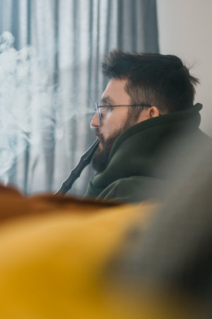 Bearded millennial or gen z man smoking hookah while relaxing on sofa at home - chill time and resting conceptの写真素材