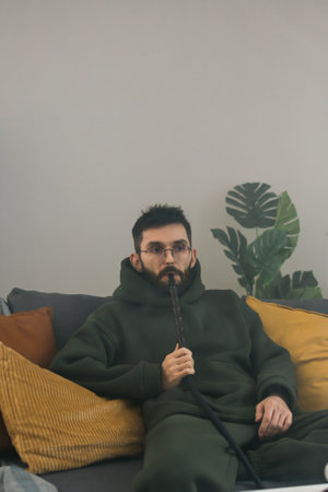 Bearded millennial or gen z man smoking hookah while relaxing on sofa at home - chill time and resting conceptの写真素材