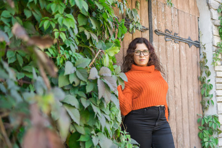 Autumn portrait of an attractive young woman in stylish glasses in a knitted fashionable orange sweater on background of leaves and big door . Girl walks in city - copy space and empty space for textの写真素材
