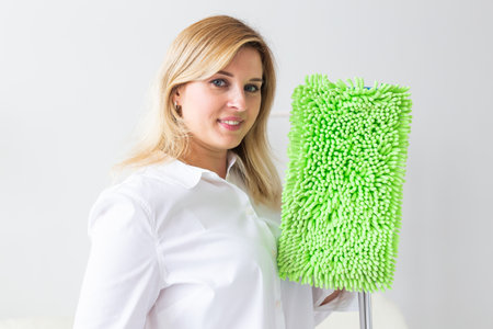 Smiling millennial woman housewife with mop for washes floor enjoys cleaning in minimalist interior....