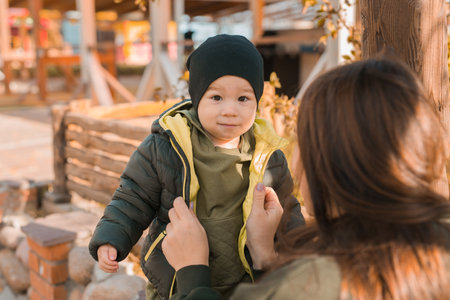 Mother with her little asian son in autumn park. Woman zips up child jacket. Fall and motherhood conceptの写真素材