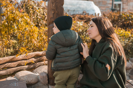 Cute little asian boy stands with mother outdoors copy space. Happy child walking in autumn park. Toddler baby boy wears trendy jacket and hat. Autumn fashion. Stylish child outside.の写真素材