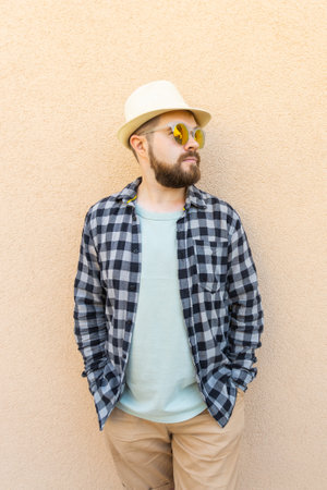 Handsome millennial man wearing summer hat and plaid shirt smiling happy near wall - travel vacations and summer holiday conceptの写真素材