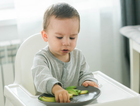 Baby boy sitting in baby chair eating fruit on kitchen background - baby feeding conceptの写真素材