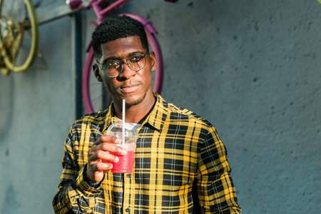 Young attractive African American hipster sipping fresh smoothie, waiting for his friends on hot summer day, hiding in the shadow. Copy spaceの写真素材