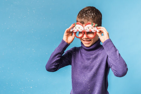 Cute little child boy in party Christmas Santa Claus glasses on blue background. Kid with snow on headの写真素材