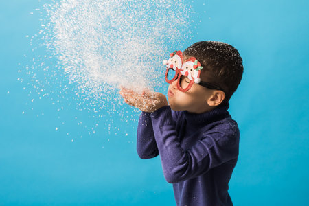 Child boy blowing snow out of his hands on blue background. Christmas and New Year holidays concept. Generation alpha and gen alpha children. Copy spaceの写真素材