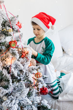 Child decorates Christmas tree. The boy is holding a Christmas toy. Morning before christmas at home. Generation alpha and gen alpha children.の写真素材