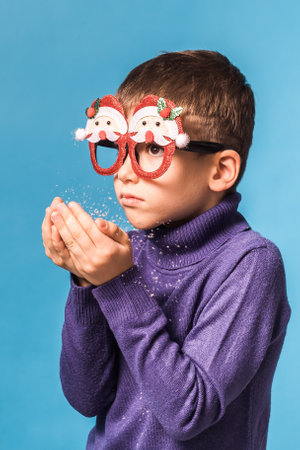 Child boy blowing snow out of his hands on blue background. Christmas and New Year holidays concept. Generation alpha and gen alpha children.の写真素材