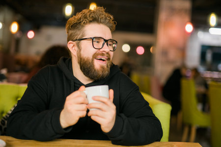 Bearded curly man smiling confident drinking coffee in restaurant or coffee shop. Millennial generation and Gen Yの写真素材
